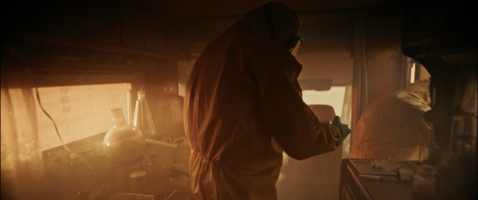 Figure in a hooded protective suit and gas mask inside a hazy, amber-lit caravan - Amon Amarth